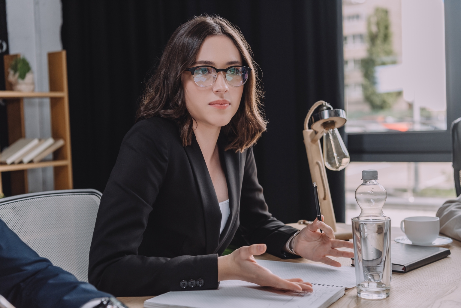 Frau mit Brille und schwarzem Blazer gestikuliert im Gespräch an einem Tisch mit Unterlagen