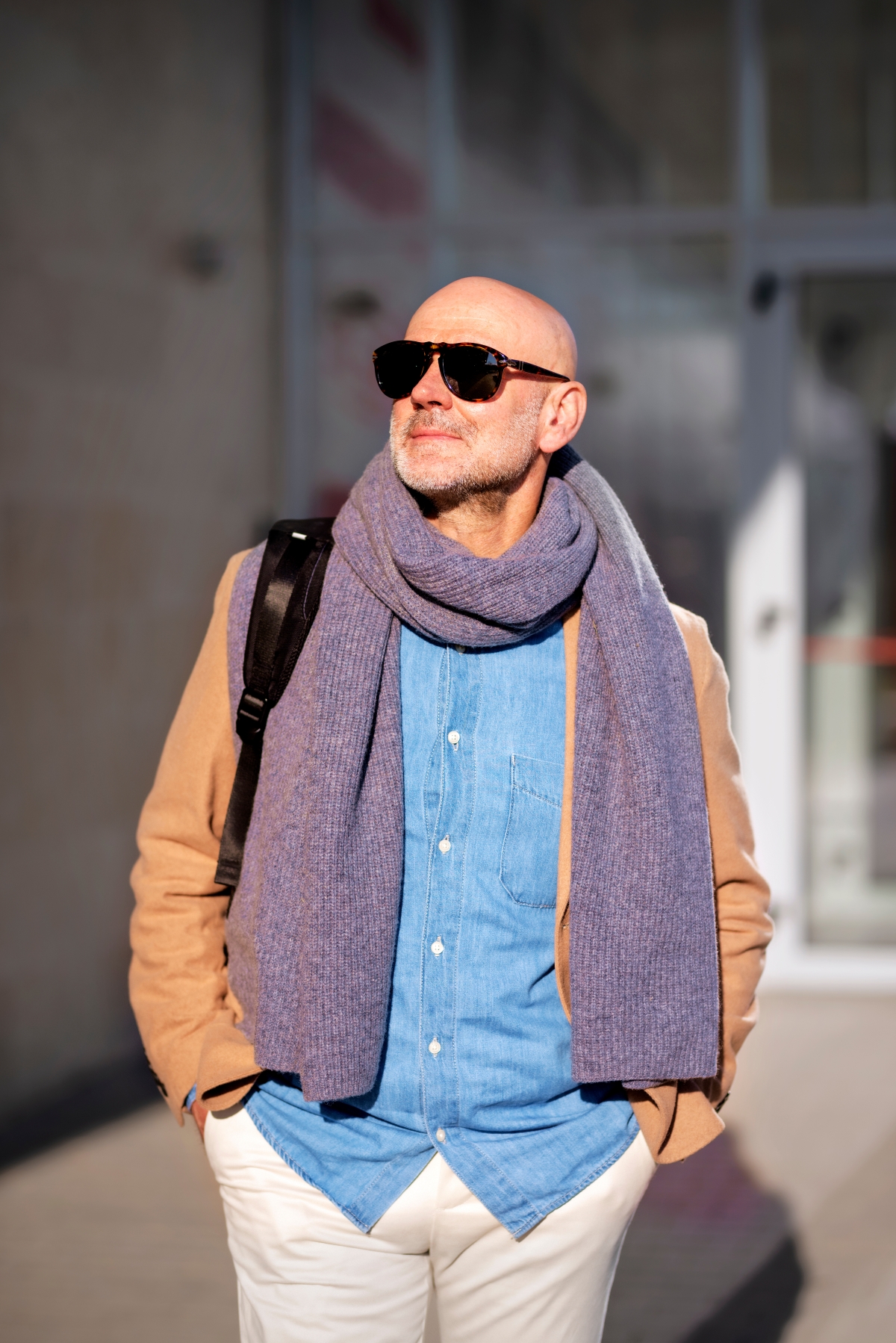 Mann mit Sonnenbrille, Schal und hellbraunem Mantel über einem Jeanshemd im Sonnenlicht.